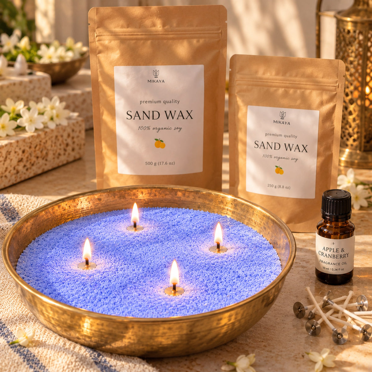 Blue sand wax candle in a brass urli with four lit wicks, beside 250 g and 500 g Mikaya sand wax pouches, fragrance oil and wicks