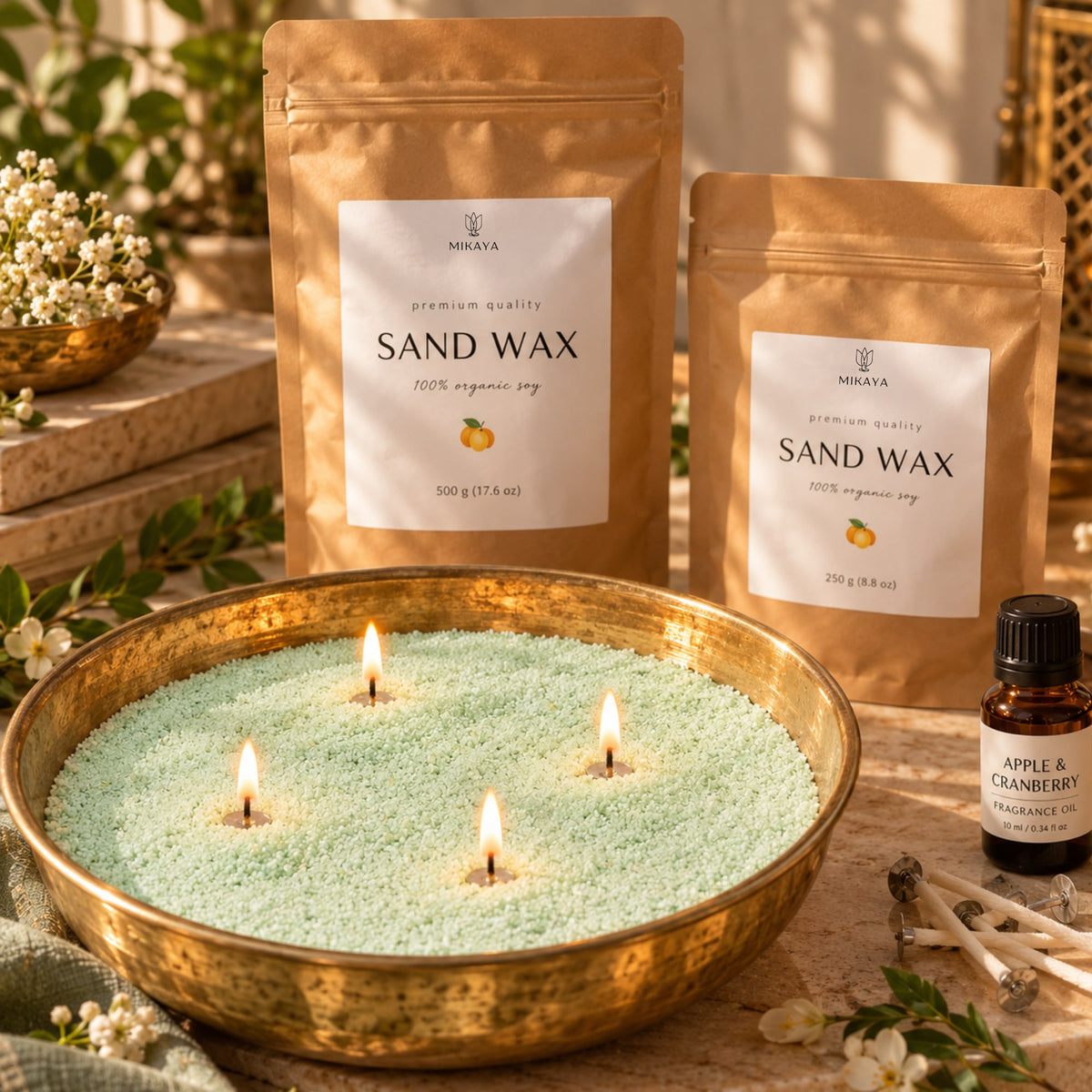 Green sand wax candle in a brass urli with four lit wicks, beside 250 g and 500 g Mikaya sand wax pouches, fragrance oil and wicks