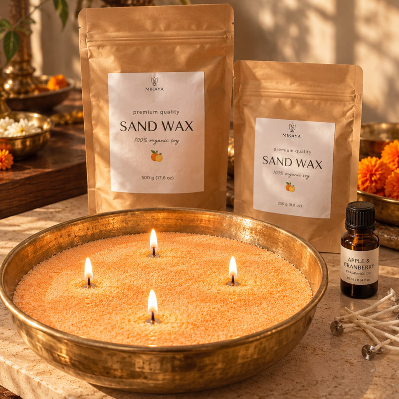 Orange sand wax candle in a brass urli with four lit wicks, beside 250 g and 500 g Mikaya sand wax pouches, fragrance oil and wicks