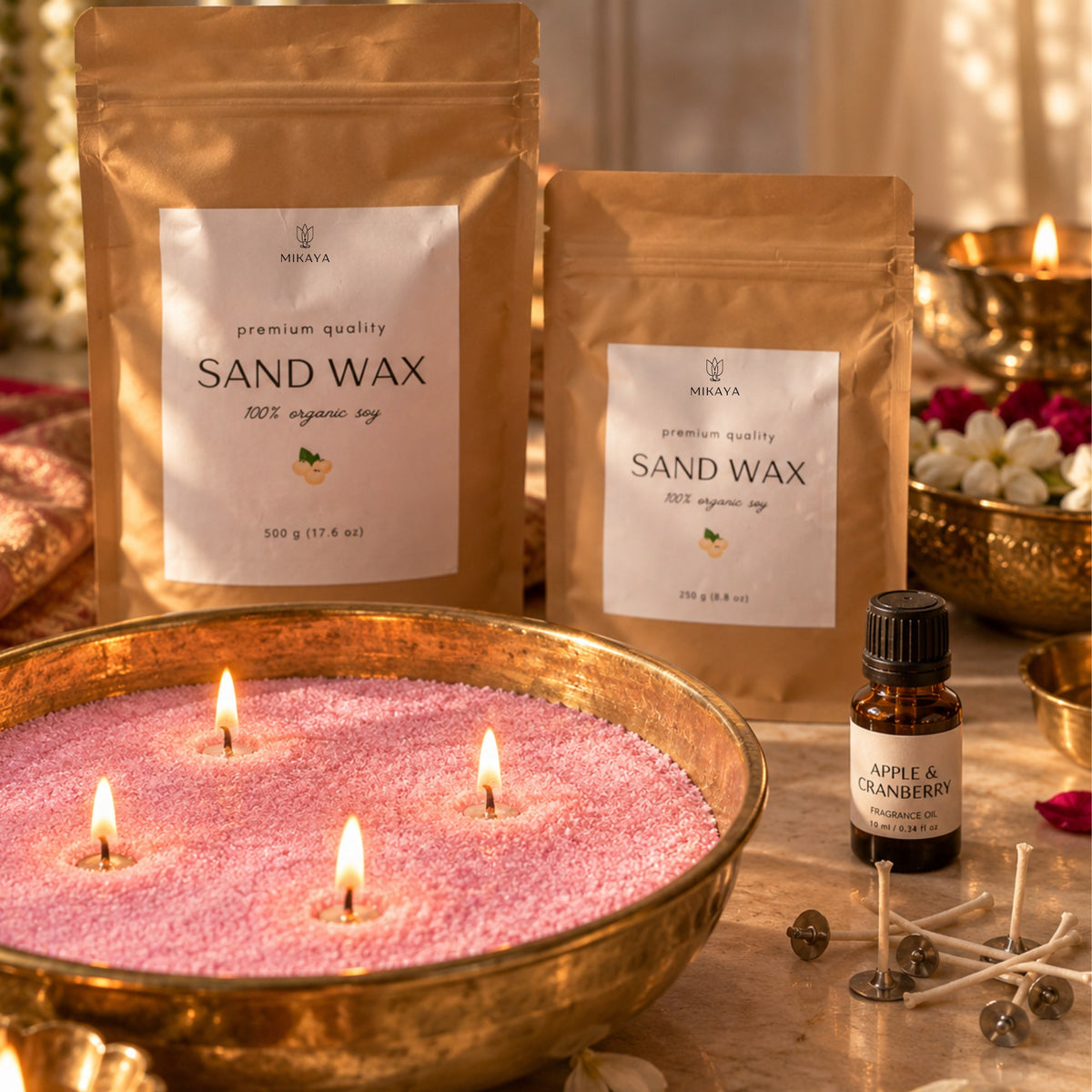 Pink sand wax candle in a brass urli with four lit wicks, beside 250 g and 500 g Mikaya sand wax pouches, fragrance oil and wicks