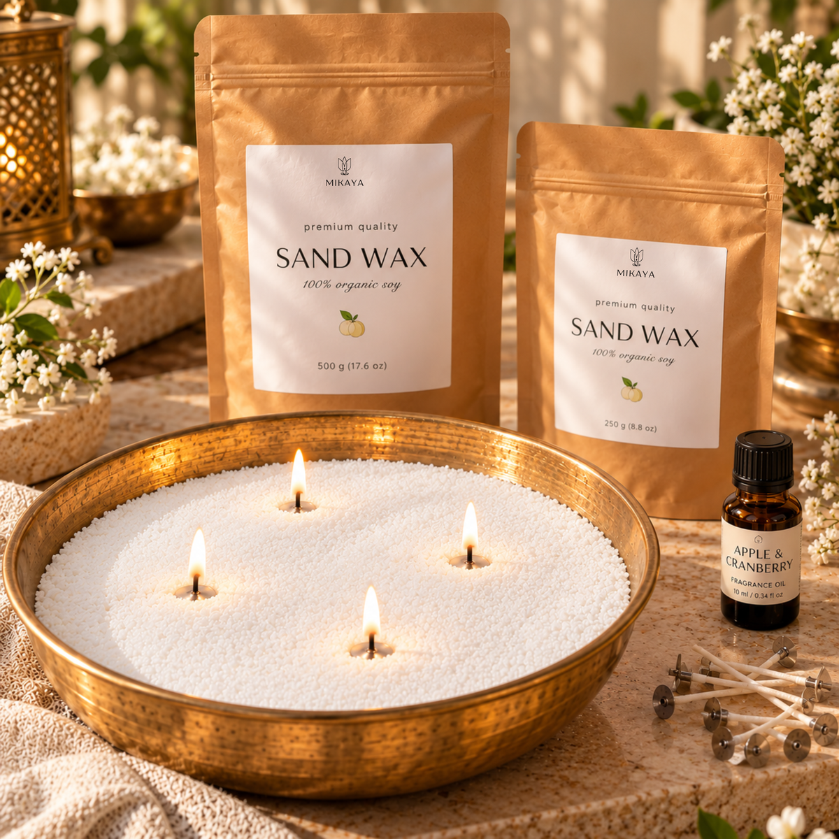 White sand wax with four lit wicks in a brass urli, beside 500 g and 250 g Mikaya sand wax pouches, a fragrance oil and cotton wicks