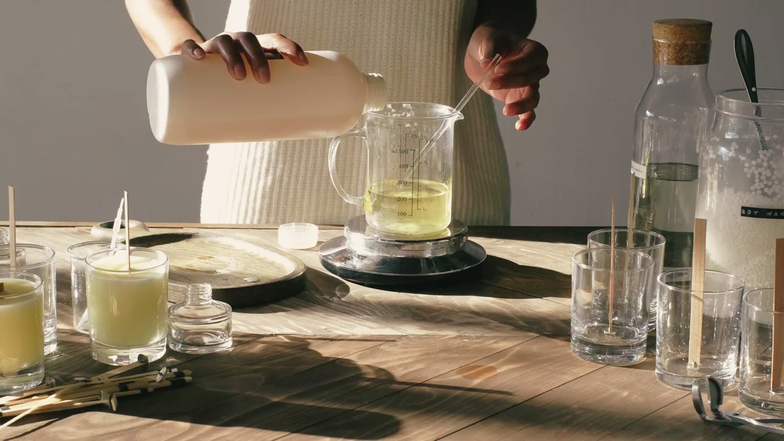 Hands pouring melted wax into glass jars with wicks set up on a wooden table before a candle making practical